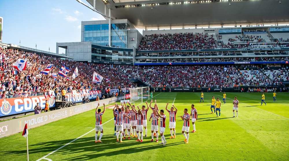 Jogadores de time com uniforme listrado em vermelho, branco e azul comemoram em campo diante de arquibancada lotada, em estádio ensolarado
