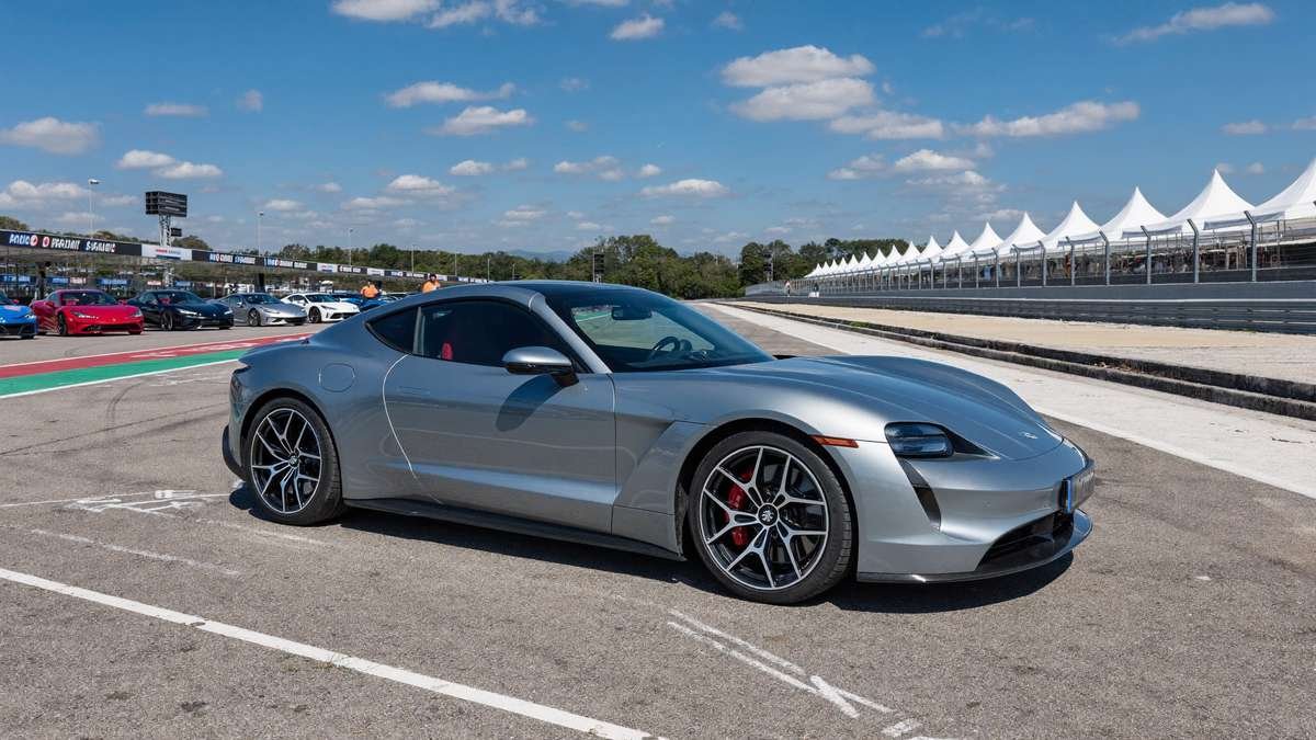 Cupê esportivo prateado em pit lane de autódromo, em dia ensolarado, com outros carros e tendas ao fundo