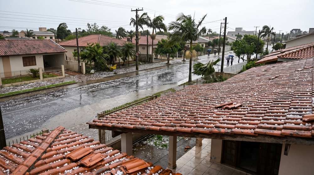Telhados de casas cobertos de granizo e rua alagada com pedestres protegidos por guarda-chuvas durante temporal em bairro residencial
