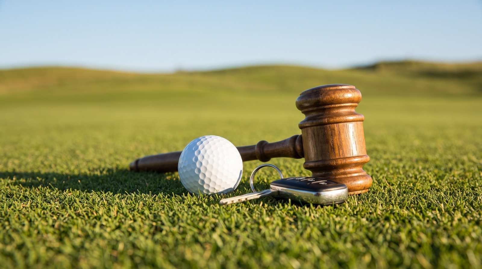 Bola de golfe, chaves de carro e um martelo de juiz sobre a grama de um campo de golfe iluminado por luz natural