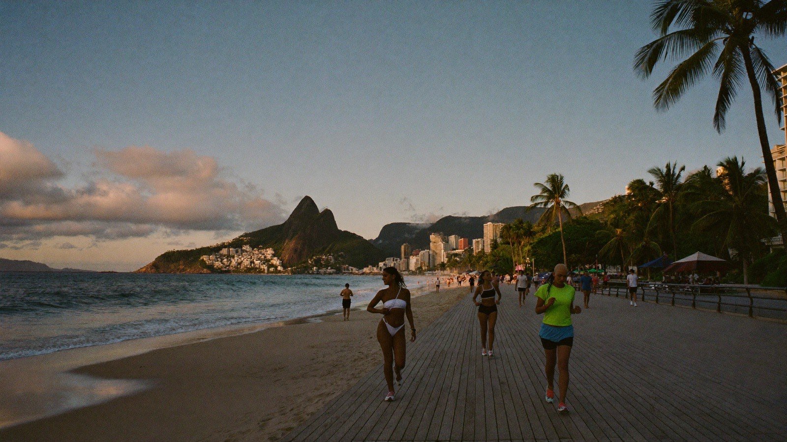 Pessoas caminhando na orla de Ipanema ao entardecer, com o morro Dois Irmãos ao fundo