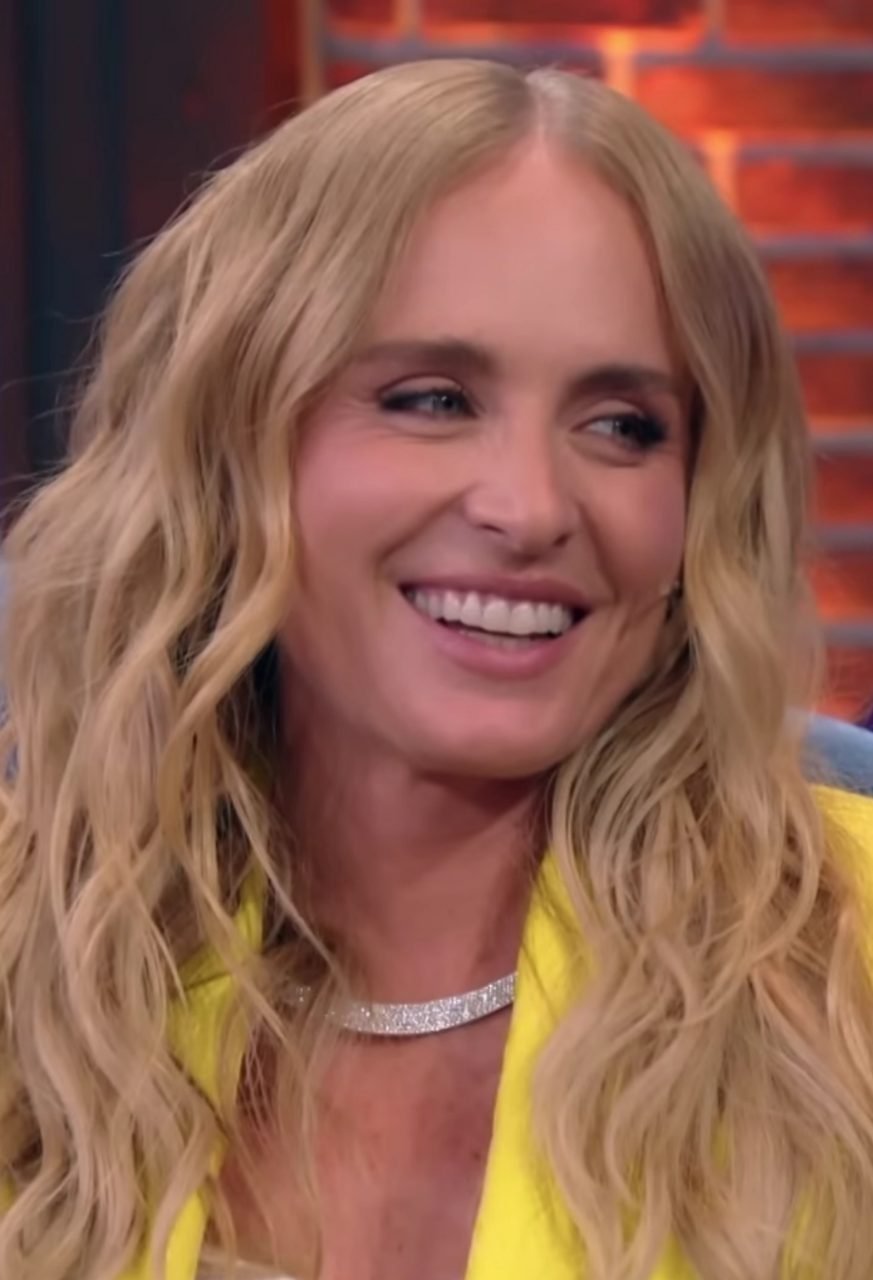 Angélica sorrindo, com cabelo loiro ondulado solto e blazer amarelo, em estúdio de televisão com iluminação quente ao fundo