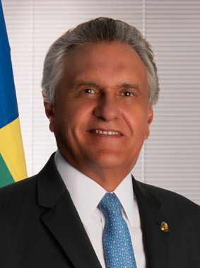 Retrato oficial de Ronaldo Caiado, de terno escuro e gravata azul, com bandeira de Goiás ao fundo