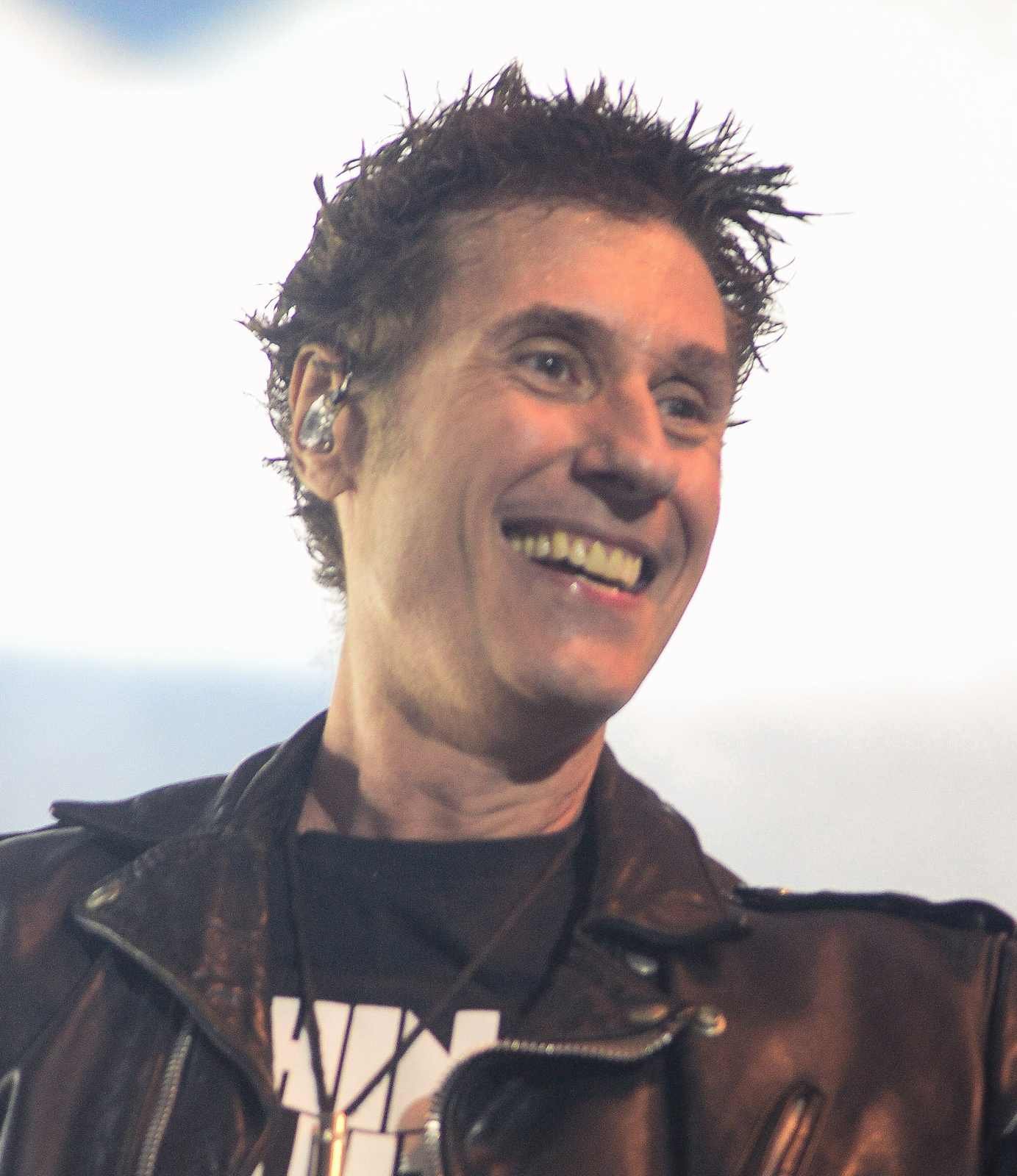 Dinho Ouro Preto, vocalista do Capital Inicial, sorrindo durante show, usando jaqueta de couro preta, com iluminação clara de palco