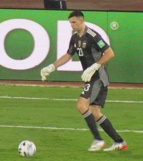 Emiliano "Dibu" Martínez, de uniforme escuro da seleção argentina, conduzindo a bola em campo bem iluminado durante partida das Eliminatórias
