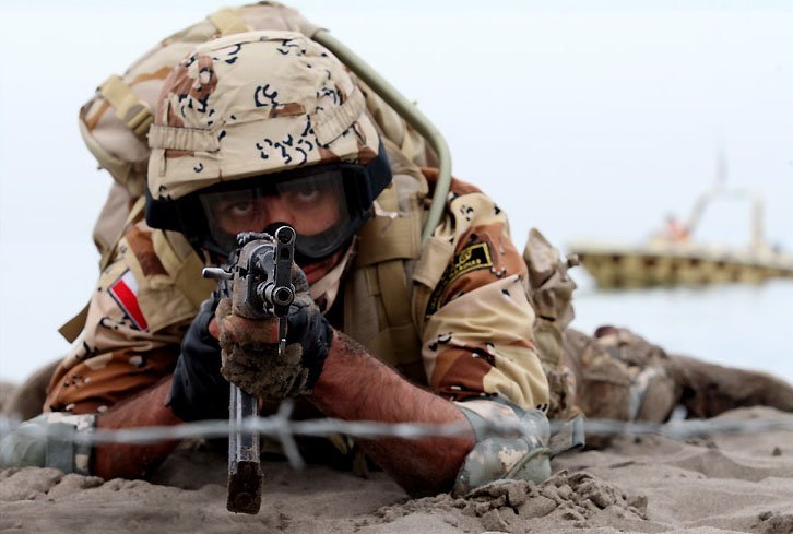 Comando naval da Guarda Revolucionária do Irã em posição de tiro na areia, com embarcação ao fundo durante exercício militar no Estreito de Ormuz