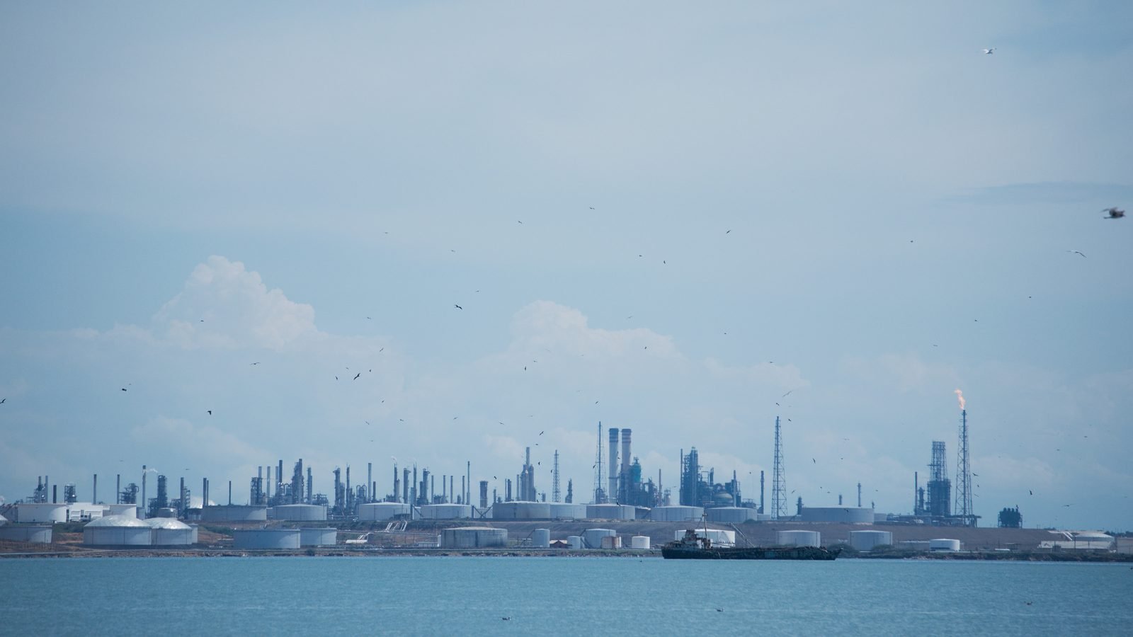 Refinaria de petróleo de Amuay, na Venezuela, vista do mar, com tanques de armazenamento, torres e uma tocha de gás acesa sob céu claro