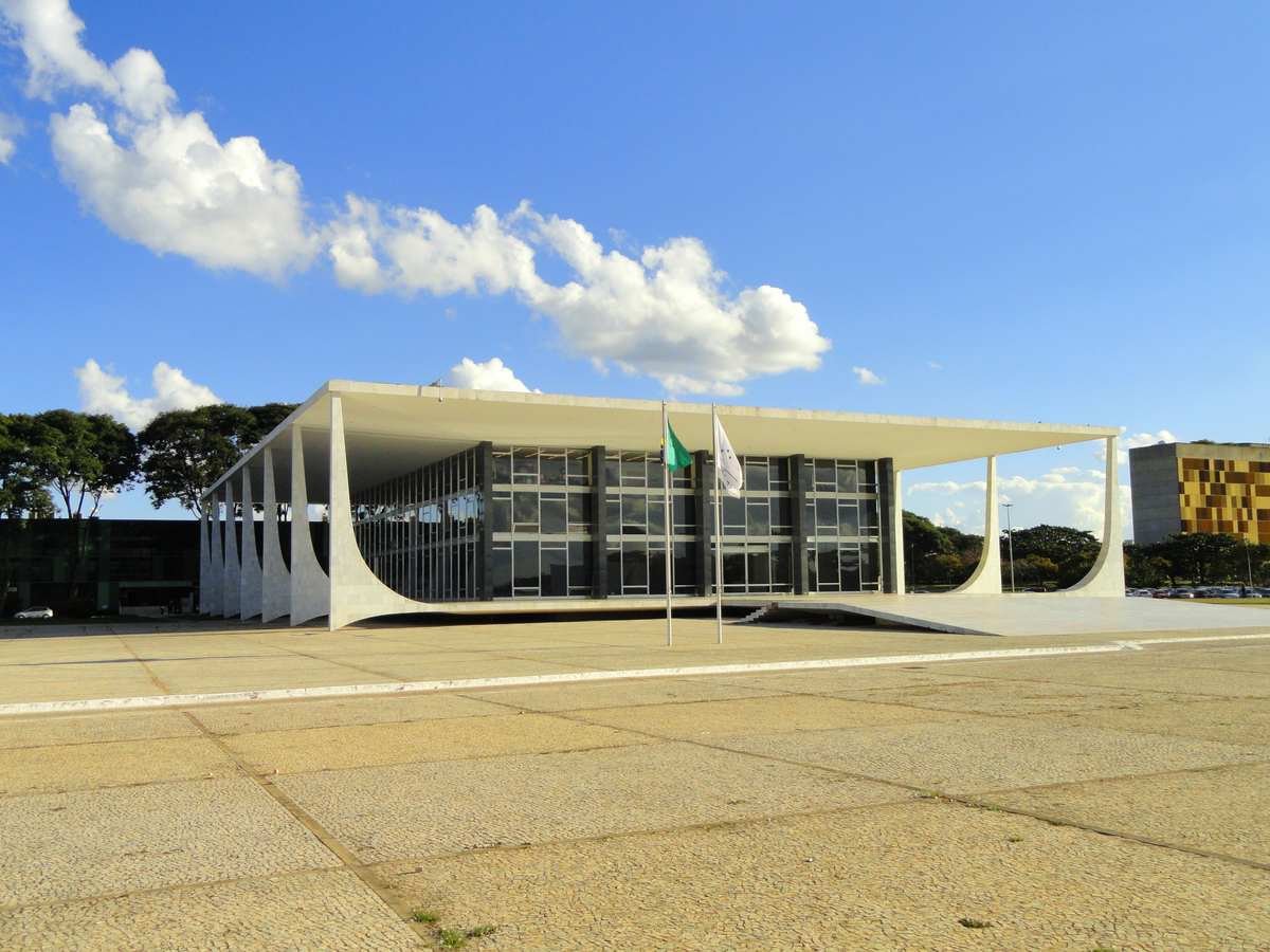 Fachada do prédio-sede do Supremo Tribunal Federal em Brasília, com colunas curvas de concreto branco, fachada de vidro e as bandeiras do Brasil e do STF hasteadas, sob céu azul de dia claro
