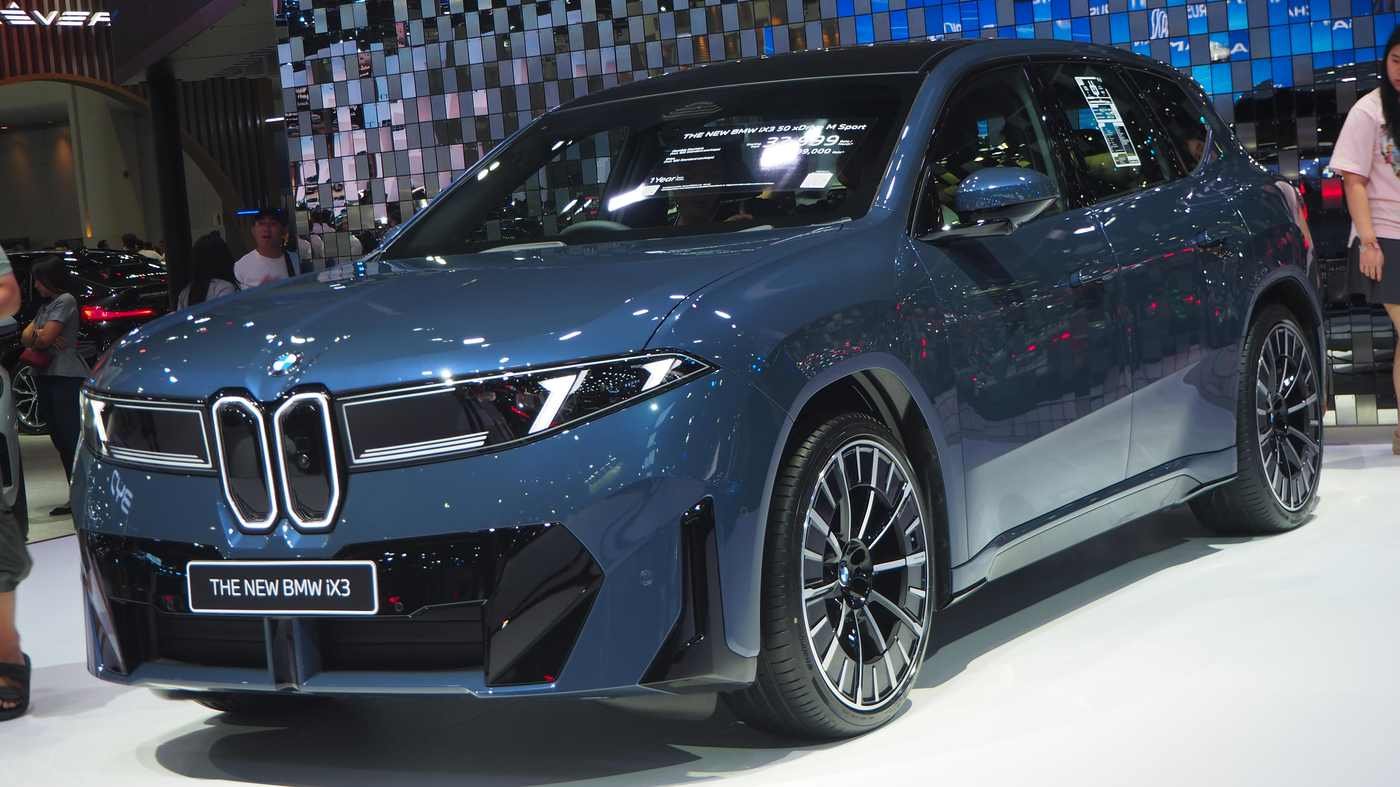 BMW iX3 50 xDrive M Sport exposto em salão automotivo, sob iluminação clara, com o logo da marca e os faróis do SUV elétrico em destaque