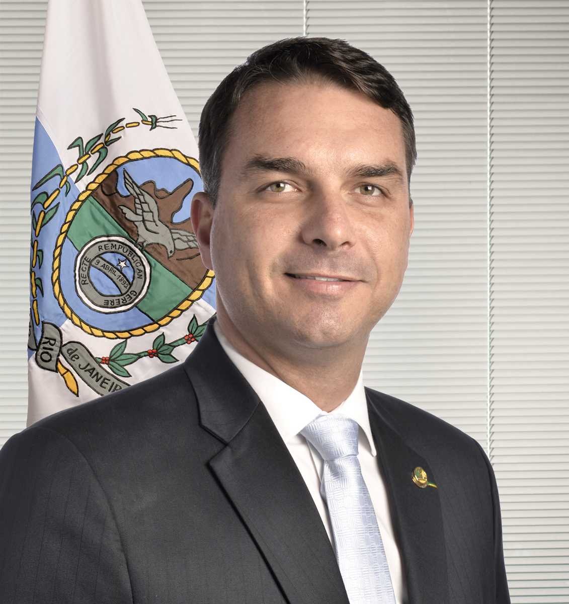Retrato oficial do senador Flávio Bolsonaro, de terno escuro e gravata clara, sorrindo diante de bandeira do Rio de Janeiro