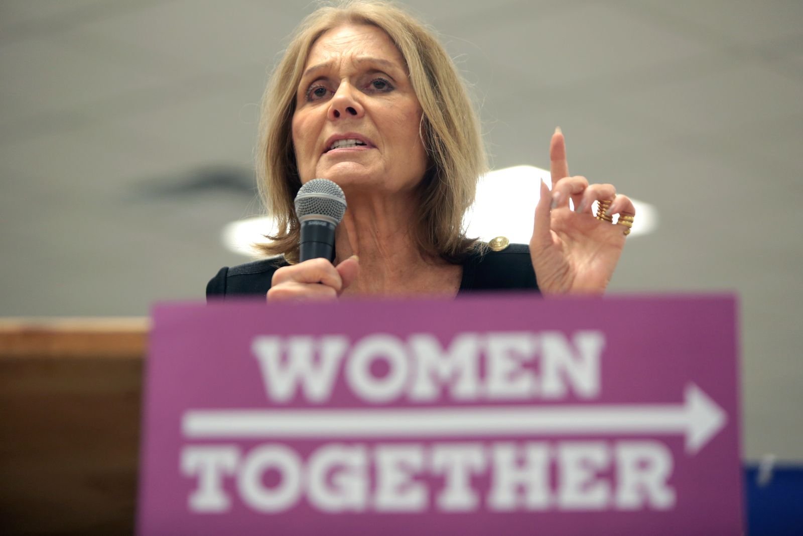 Gloria Steinem discursa ao microfone em evento pelos direitos das mulheres, com cartaz "Women Together" ao fundo