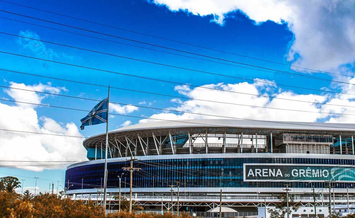 Fachada da Arena do Grêmio em dia de céu azul, com a bandeira do clube hasteada e o letreiro "Arena do Grêmio" iluminado pelo sol