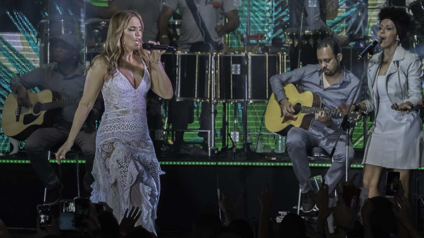 Ivete Sangalo cantando no palco com a banda ao fundo e o público de braços erguidos