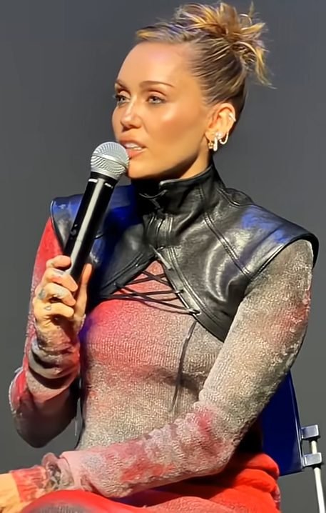 Miley Cyrus fala ao microfone durante evento com jaqueta de couro preta e coque no cabelo