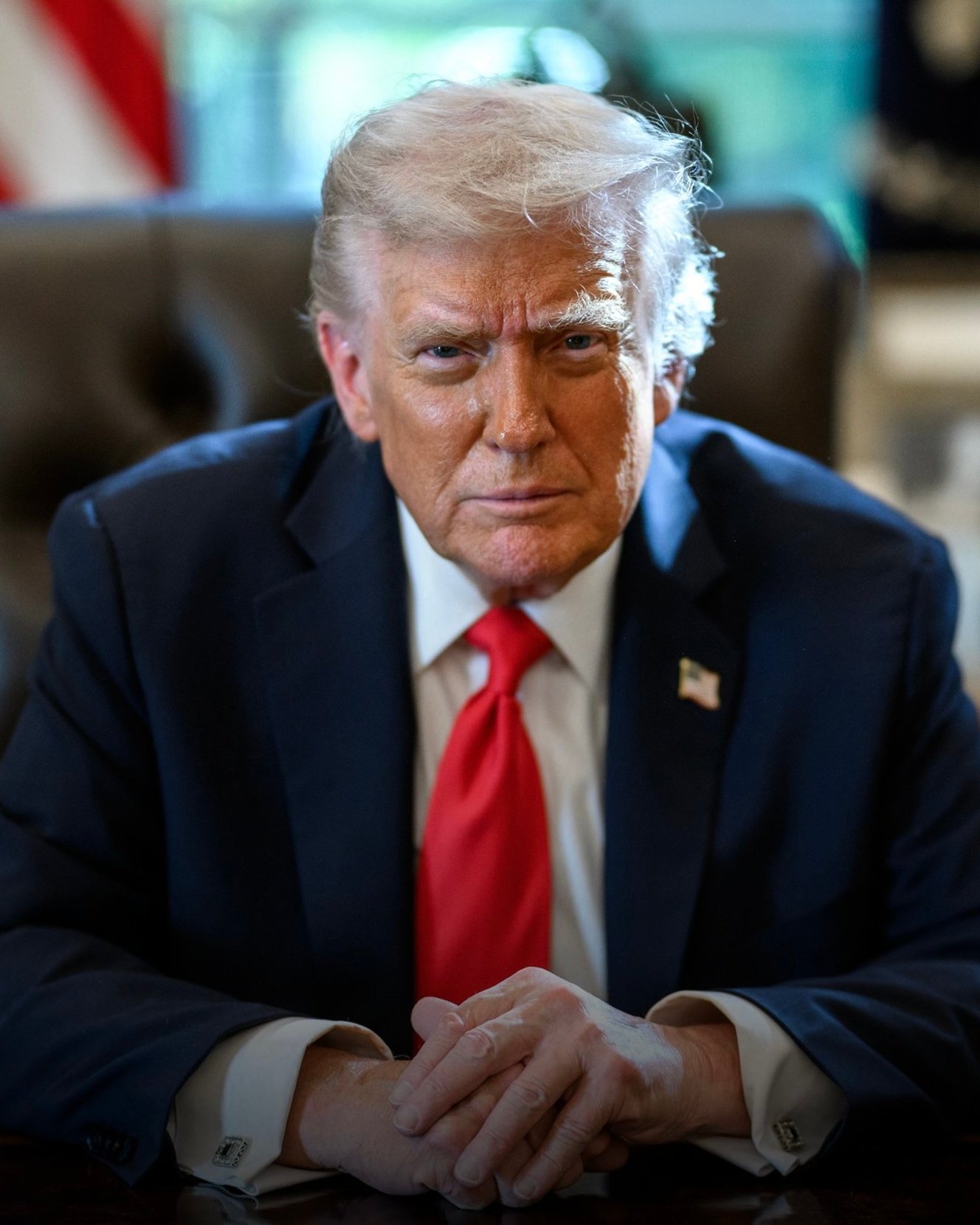 Donald Trump sentado, de terno azul-marinho e gravata vermelha, com as mãos entrelaçadas sobre uma mesa