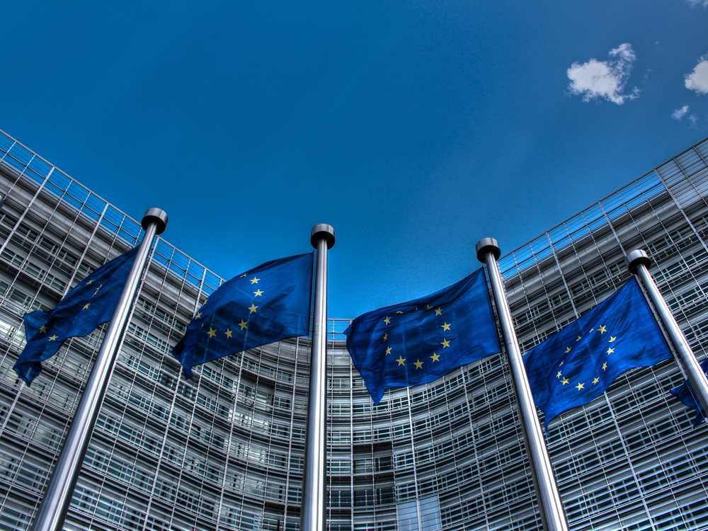 Bandeiras da União Europeia hasteadas em frente ao edifício Berlaymont, sede da Comissão Europeia em Bruxelas, sob céu azul e ensolarado