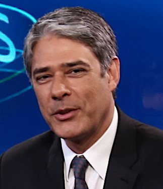 William Bonner sorrindo em estúdio de televisão com fundo azul, usando terno escuro, camisa branca e gravata